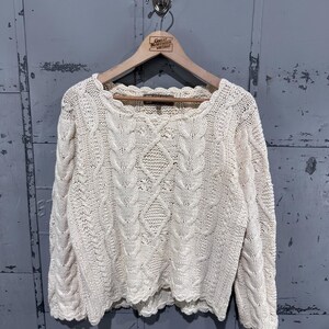 May include: An off-white long-sleeve sweater featuring a prominent cable knit pattern with vertical braids and diamond shapes. The garment has a scalloped edge along the neckline, cuffs, and hem, and is displayed on a wooden hanger.