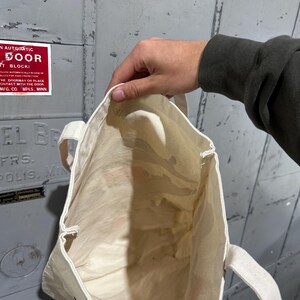May include: A cream-colored tote bag with matching handles is held open, revealing the interior. The bag appears to be made of a lightweight fabric. The background is a gray metal door with a red sign that reads "IN AUTOMATIC DOOR".