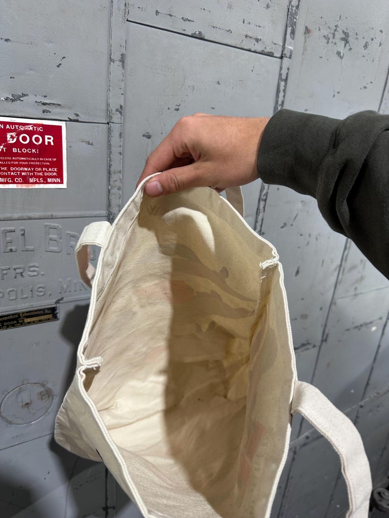 May include: A cream-colored tote bag with matching handles is held open, revealing the interior. The bag appears to be made of a lightweight fabric. The background is a gray metal door with a red sign that reads "IN AUTOMATIC DOOR".