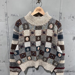 May include: A long-sleeve knit jumper featuring a high mock neck and a chequered pattern in shades of grey, brown, and blue. The centre panel displays a grid of squares with small embroidered floral accents, while the sleeves feature horizontal stripes.