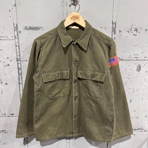 May include: An olive green button-up shirt with two chest pockets and an American flag patch on the sleeve. The shirt is hanging on a wooden hanger. The shirt has a collared neckline and button closures.