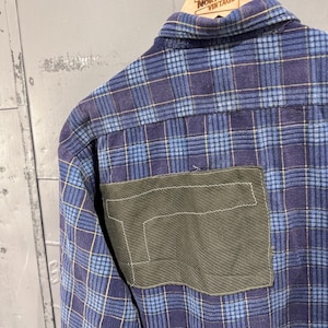 Pu&ograve; includere: Una camicia in flanella a quadri blu e viola con una grande toppa rettangolare in tessuto verde oliva cucita sul retro. La camicia presenta un colletto standard ed &egrave; appesa a una gruccia in legno con la scritta Great Northern Vintage.