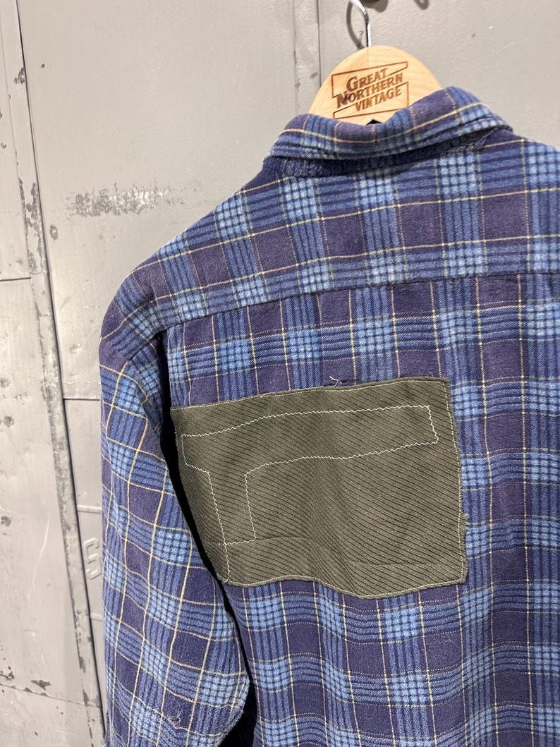 Pu&ograve; includere: Una camicia in flanella a quadri blu e viola con una grande toppa rettangolare in tessuto verde oliva cucita sul retro. La camicia presenta un colletto standard ed &egrave; appesa a una gruccia in legno con la scritta Great Northern Vintage.