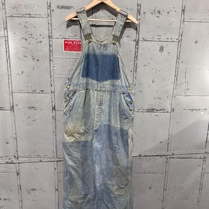 Può includere: Salopette in denim blu chiaro con lavaggio sbiadito e tasca a pettorina. La salopette ha spalline regolabili, elementi metallici e un classico design da lavoro. L'indumento è appeso a una gruccia di legno.