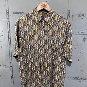 May include: Short-sleeved shirt with a geometric pattern in tan, navy, and cream. The shirt is hanging on a wooden hanger with the text "Great Northern Vintage". The shirt has a chest pocket.