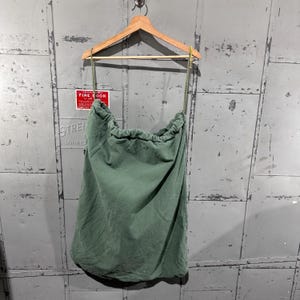 以下が含まれることがあります： 木製のハンガーに吊るされた、巾着開閉式の緑色のキャンバスランドリーバッグ。 バッグはオリーブグリーン一色で、丈夫な素材でできているようです。 背景は風化した金属面です。