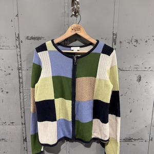 May include: A patchwork zip-up cardigan in a variety of colors, including green, blue, beige, and cream. The sweater is hanging on a wooden hanger. The cardigan has a navy blue trim and a zipper closure.