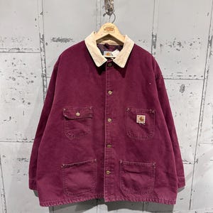 May include: A burgundy work jacket with a cream-colored corduroy collar. The jacket has four front pockets, button closures, and a Carhartt logo patch. The jacket is hanging on a wooden hanger.