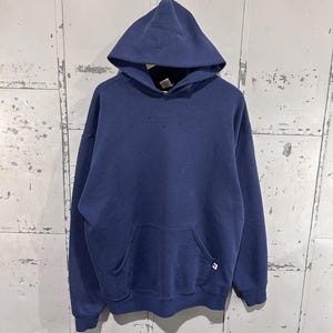 May include: A navy blue hooded sweatshirt with a front pocket and a drawstring hood. The sweatshirt has a small logo on the lower right side. The garment is hanging against a weathered, metallic background.
