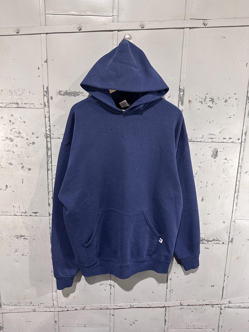 May include: A navy blue hooded sweatshirt with a front pocket and a drawstring hood. The sweatshirt has a small logo on the lower right side. The garment is hanging against a weathered, metallic background.