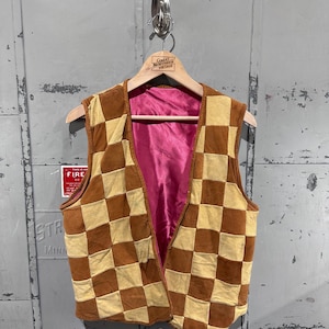 May include: A vintage, open-front vest with a checkerboard pattern in shades of brown and tan. The vest has a pink satin lining and is hanging on a wooden hanger. The hanger has a label that reads "Great Northern Vintage".