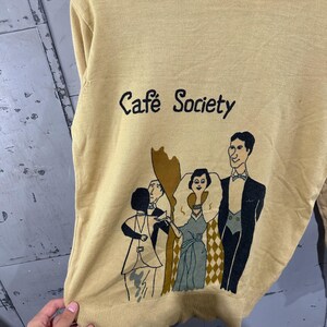 May include: Mustard yellow sweater with the text "Caf&eacute; Society" and a graphic of people in formal attire. The illustration features figures in black, white, and shades of green and brown. The sweater has a ribbed neckline and hem.