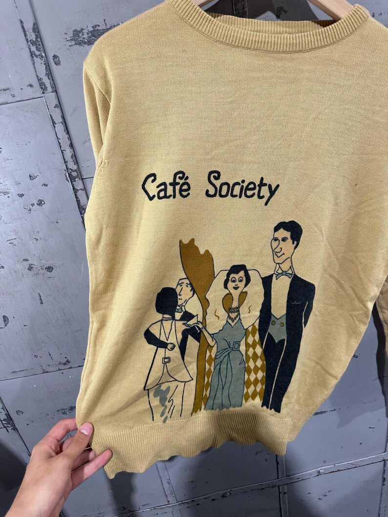 May include: Mustard yellow sweater with the text "Caf&eacute; Society" and a graphic of people in formal attire. The illustration features figures in black, white, and shades of green and brown. The sweater has a ribbed neckline and hem.