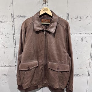 May include: Brown leather jacket with a zip-up front, two front pockets, and a ribbed hem and cuffs. The jacket is hanging on a wooden hanger with the text "Great Northern Vintage".