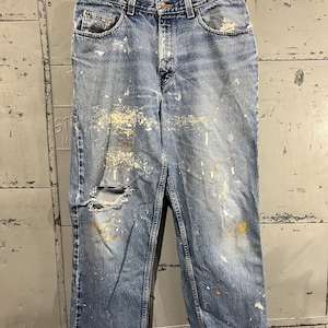 May include: Distressed blue denim jeans with paint splatters and a small hole on the left leg. The jeans have a faded look and are worn.