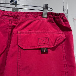 May include: Red drawstring shorts with a black pocket and a black and orange drawstring. The pocket has a black X stitched on it.