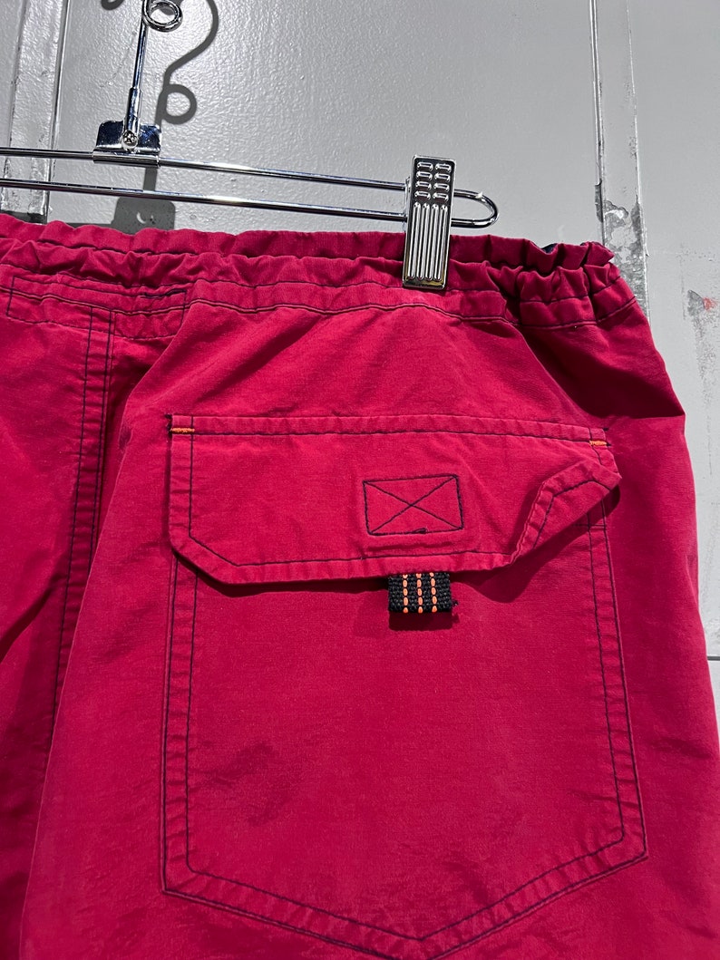 May include: Red drawstring shorts with a black pocket and a black and orange drawstring. The pocket has a black X stitched on it.