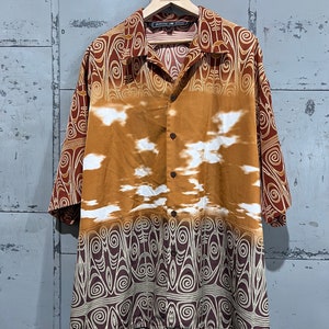 May include: A brown and white short-sleeved button-down shirt with a geometric pattern. The shirt has a collar and a button-up front. The pattern is a mix of brown, white, and gold.