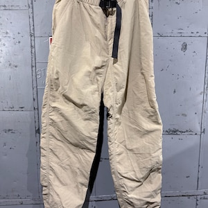 May include: Tan-colored pants with an elastic waistband and a black belt. The pants have a relaxed fit with gathered ankles. The fabric appears lightweight and suitable for casual wear. The pants are hanging on a wooden hanger.