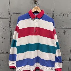 Può includere: Una maglia da rugby a maniche lunghe con strisce orizzontali rosse, bianche, turchesi e blu. La maglia ha colletto e polsini rossi e una paramontura a tre bottoni. La maglia è appesa a una gruccia di legno.