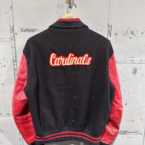 Op de afbeelding: Zwarte en rode varsity jas met het woord "Cardinals" in een cr&egrave;mekleurig script op de rug. De jas heeft rode leren mouwen en een zwarte body. De kraag en manchetten hebben rood-zwarte strepen.