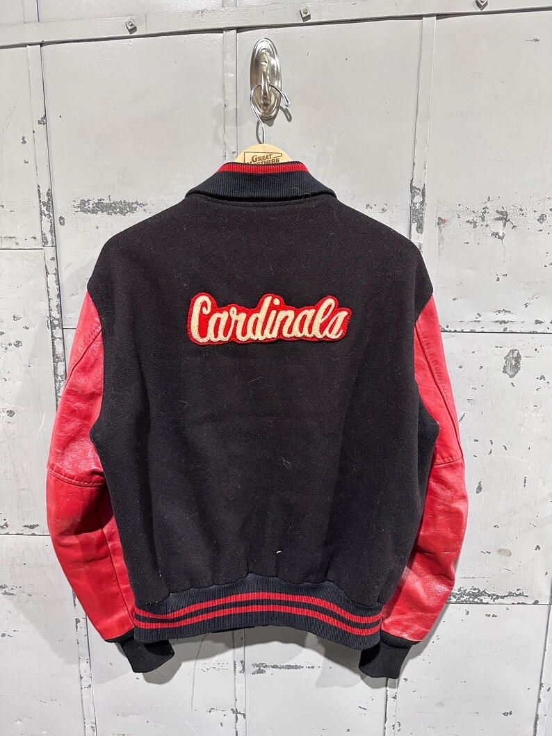 Op de afbeelding: Zwarte en rode varsity jas met het woord "Cardinals" in een cr&egrave;mekleurig script op de rug. De jas heeft rode leren mouwen en een zwarte body. De kraag en manchetten hebben rood-zwarte strepen.