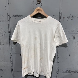 Könnte beinhalten: Ein schlichtes, cremeweißes T-Shirt mit kurzen Ärmeln und Rundhalsausschnitt. Der Stoff weist sichtbare hellbraune Flecken und Verfärbungen auf der unteren linken Seite und in der Mitte auf, was auf Gebrauchsspuren und Alter hinweist.