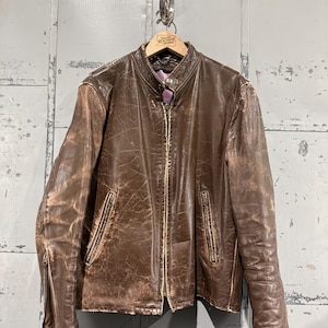 May include: A brown leather jacket with a zipper closure and two front pockets. The jacket has a worn, distressed appearance, with visible wear and tear on the leather. It hangs on a wooden hanger.