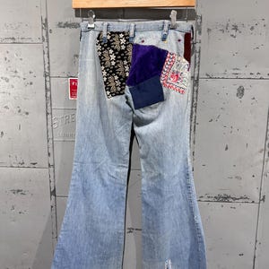 May include: Light blue denim jeans with a flared leg and patches on the back pockets. The patches feature floral, paisley, and solid fabrics. The jeans have a raw hem and are hanging on a wooden hanger. The jeans are approximately 102 cm in length.