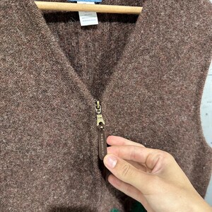 K&ouml;nnte beinhalten: Eine braune Weste aus Wolle mit Frontrei&szlig;verschluss und einem Lederanh&auml;nger, der mit dem Pendleton-Logo gepr&auml;gt ist. Das Kleidungsst&uuml;ck verf&uuml;gt &uuml;ber ein Pendleton Originals-Etikett am Halsausschnitt und einen strukturierten Strickstoff mit gr&uuml;nen Baumapplikationen.