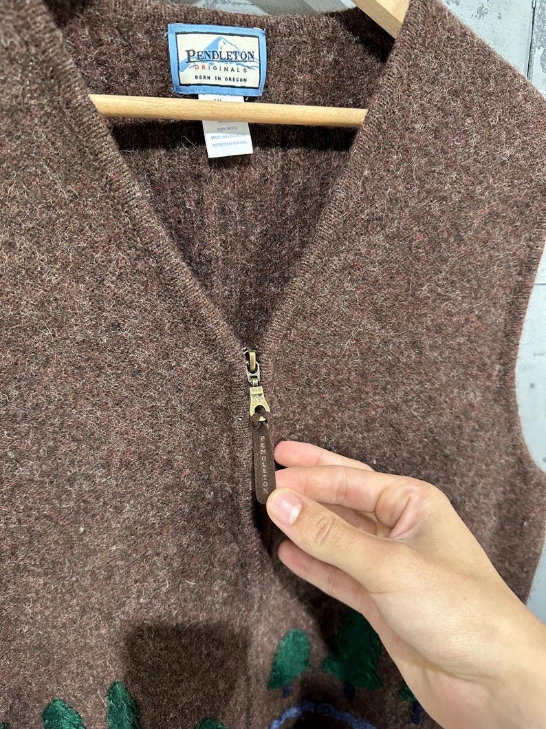 K&ouml;nnte beinhalten: Eine braune Weste aus Wolle mit Frontrei&szlig;verschluss und einem Lederanh&auml;nger, der mit dem Pendleton-Logo gepr&auml;gt ist. Das Kleidungsst&uuml;ck verf&uuml;gt &uuml;ber ein Pendleton Originals-Etikett am Halsausschnitt und einen strukturierten Strickstoff mit gr&uuml;nen Baumapplikationen.