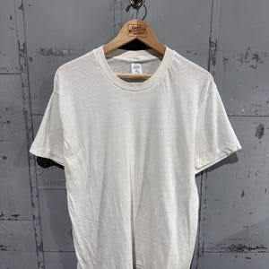 May include: A plain, off-white short-sleeve t-shirt with a crew neckline. The shirt is made of a solid, light-colored fabric and features a standard fit with short sleeves and a straight hemline. A small white tag is visible at the inner collar.