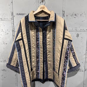 May include: A quilted jacket with a beige base, vertical black and floral patterned stripes, and light blue accents. The jacket has a collared neckline, two front pockets, and is hanging on a wooden hanger.