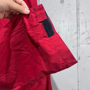 May include: Close-up of the red fabric of a pair of trousers with a black adjustable strap on the side seam.
