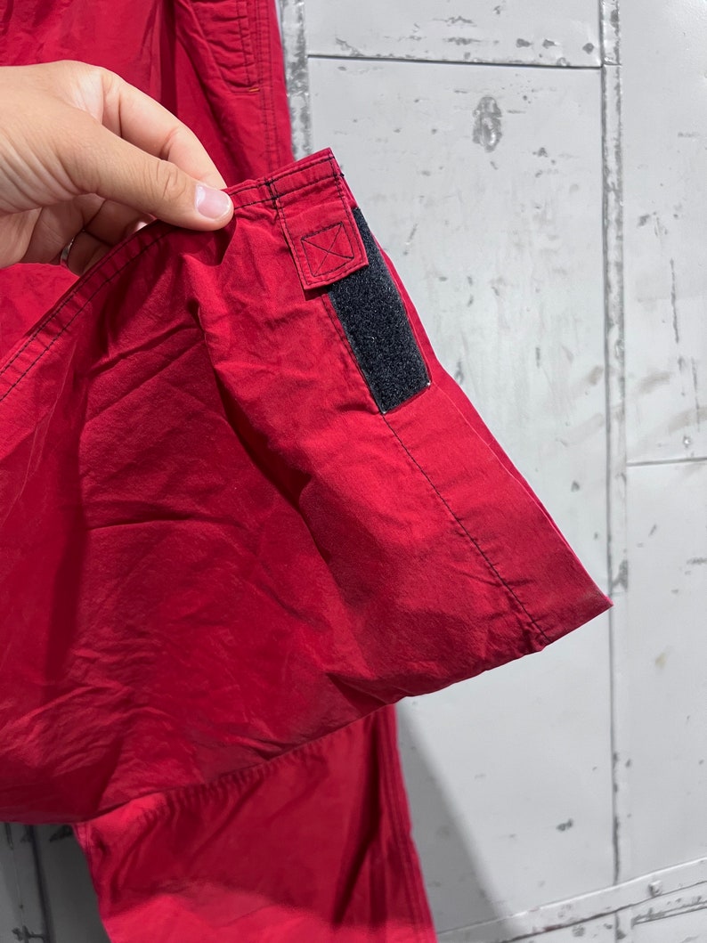 May include: Close-up of the red fabric of a pair of trousers with a black adjustable strap on the side seam.