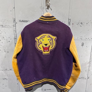 Op de afbeelding: Een vintage paarse en gouden varsity jas met een tijgerembleem op de rug. De jas heeft gele leren mouwen en gestreepte details op de kraag, manchetten en zoom. Het tijgerembleem is geel met groene ogen en een rode mond.