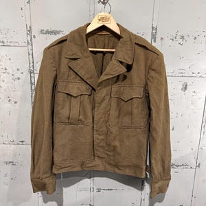 May include: A brown wool military-style jacket featuring a collared neckline, long sleeves with buttoned cuffs, and two large chest pockets with pointed flaps. The jacket has shoulder epaulets and a straight hemline, providing a structured fit.