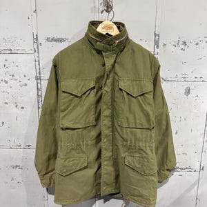 May include: Olive green military-style jacket with multiple front pockets and a high collar. The jacket has a zipper closure and a drawstring at the waist. The jacket is hanging on a metal hook against a weathered, gray background.