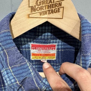 Pu&ograve; includere: Una camicia di flanella a quadri blu con colletto e un'etichetta bianca con il testo FLANNELCRAFT, RN 35588, 100% COTTON e STYLE NO. 454P. La camicia &egrave; esposta su una gruccia di legno con il testo GREAT NORTHERN VINTAGE.