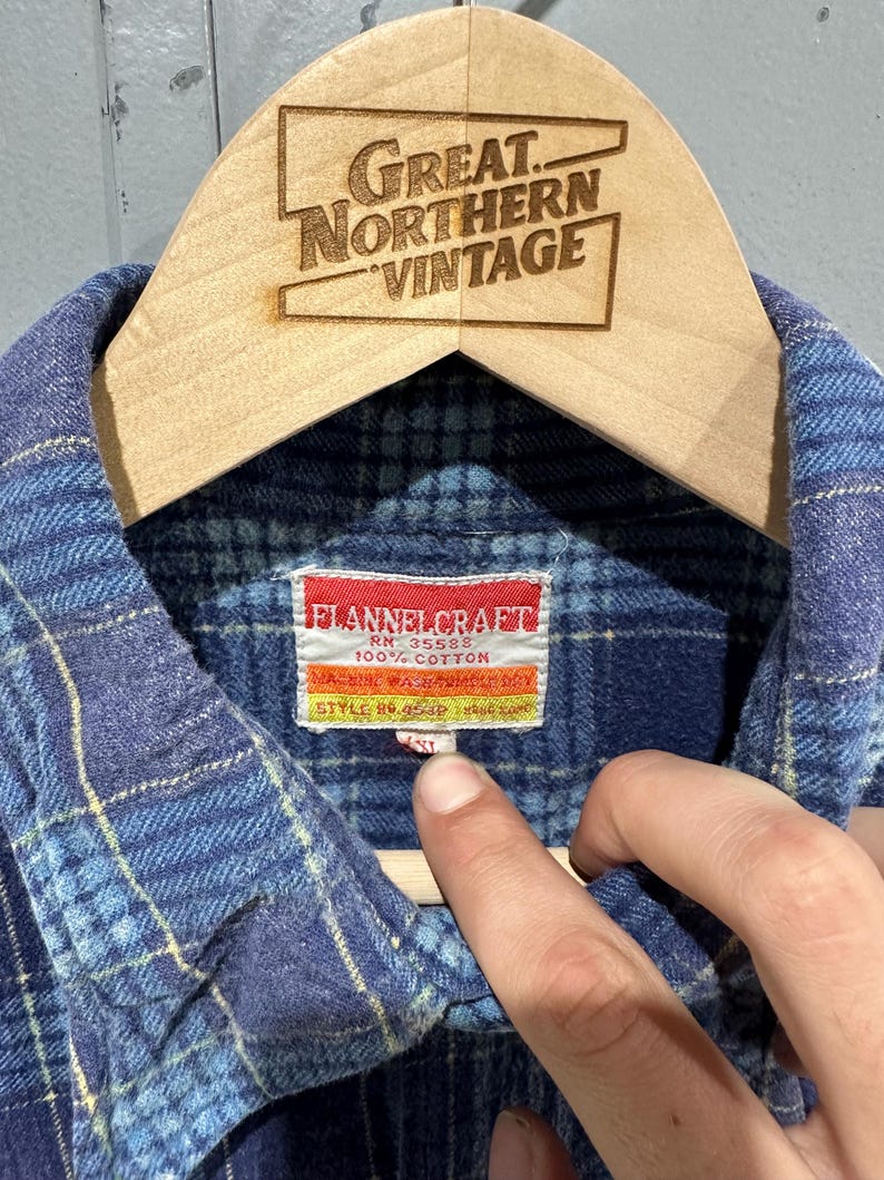 Pu&ograve; includere: Una camicia di flanella a quadri blu con colletto e un'etichetta bianca con il testo FLANNELCRAFT, RN 35588, 100% COTTON e STYLE NO. 454P. La camicia &egrave; esposta su una gruccia di legno con il testo GREAT NORTHERN VINTAGE.