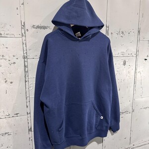 May include: A navy blue hooded sweatshirt with a front pocket and a drawstring hood. The sweatshirt has a small white and blue logo on the front pocket. The garment is hanging against a weathered metal background.