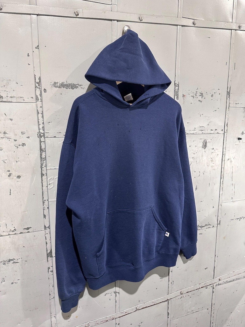 May include: A navy blue hooded sweatshirt with a front pocket and a drawstring hood. The sweatshirt has a small white and blue logo on the front pocket. The garment is hanging against a weathered metal background.