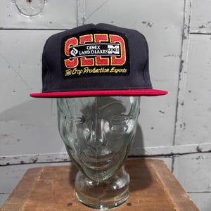Puede incluir: Gorra gris oscura de ala plana con visera roja. La gorra presenta un parche con el texto "CENEX LAND O LAKES CENEX" en rojo y blanco, y las palabras "The Crop Production Experts" están impresas debajo.
