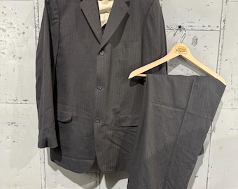 May include: A charcoal gray suit, including a blazer and trousers, hangs on wooden hangers against a weathered, metallic background. The blazer has three buttons and a notched lapel. The trousers are neatly folded below.