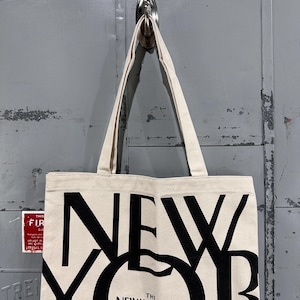 May include: A cream-colored canvas tote bag featuring large, bold black lettering that spells out NEW YORKER. The design includes the smaller text THE NEW YORKER centered within the letter O. The bag has two matching fabric shoulder straps.