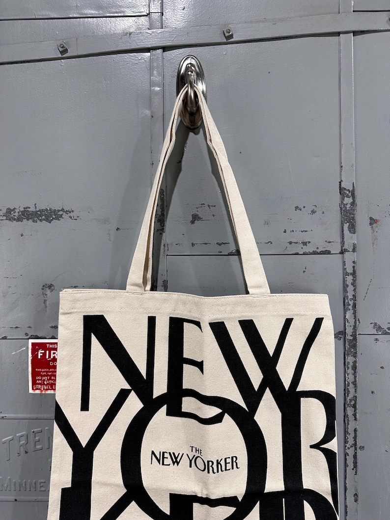 May include: A cream-colored canvas tote bag featuring large, bold black lettering that spells out NEW YORKER. The design includes the smaller text THE NEW YORKER centered within the letter O. The bag has two matching fabric shoulder straps.