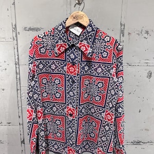 Könnte beinhalten: Ein langärmliges Hemd mit geometrischem Muster in Rot, Marineblau und Weiß, das an einem Holzbügel hängt. Das Muster wiederholt florale und geometrische Formen. Das Hemd hat eine Knopfleiste.