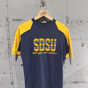 Peut inclure: T-shirt bleu marine et jaune à manches courtes. Le t-shirt présente des manches jaunes et le texte "SDSU" dans une police stylisée, avec "SOUTH DAKOTA STATE UNIVERSITY" en dessous. Le t-shirt est suspendu à un cintre en bois.