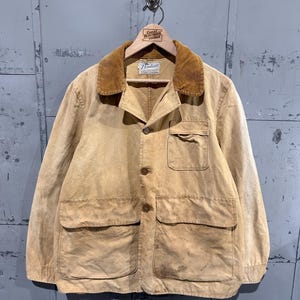 May include: A tan canvas chore coat featuring a brown corduroy collar, a button-front closure, and three front pockets. The interior label reads Pontiac Sportswear. The jacket has long sleeves and a straight hem, displaying a worn, weathered texture.