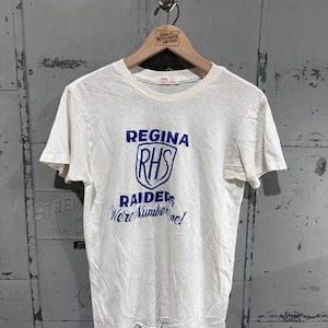 May include: Cream-colored t-shirt with the text "Regina RHS Raiders We're Number One!" in blue. The design features a shield graphic. The shirt is hanging on a wooden hanger.